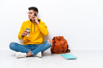 Young student caucasian man sitting one the floor isolated on white background holding coffee to take away and a mobile