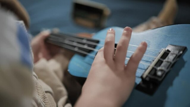 Top view of right hand of small child pulls strings on ukulele. Kid playing ukulele