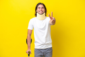 Young handsome man wearing neck brace and crutches isolated on yellow background smiling and...
