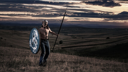 Man with bare torso in helmet with spear and shield in his hands poses in front of camera in valley