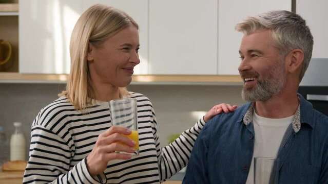 Happy couple family Caucasian adult woman and mature man middle-aged husband and wife drinking orange juice in home kitchen laughing enjoy tasty healthy fruit fresh drink laugh smiling diet healthcare