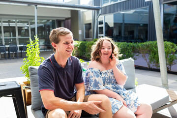 Teenager talking with young adult disability worker outside at café sitting on lounge together