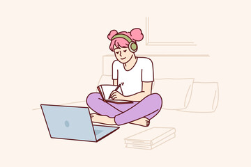 Schoolgirl watches webinar in laptop receiving distance education sitting on bed and making notes in workbook. Girl is engaged in self-education watching webinars and educational videos 