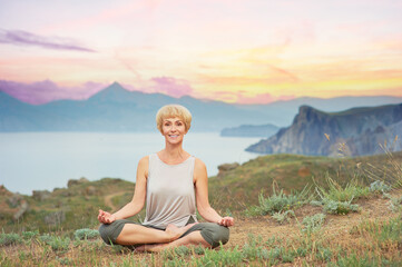 Naklejka na ściany i meble Senior woman doing yoga exercises with mountain sunset on the background