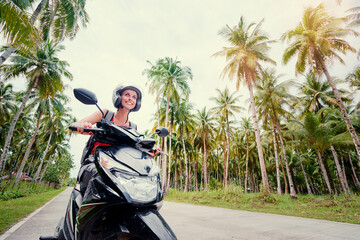 Tropical travel and transport. Young beautiful woman in helmet riding scooter on the road with palm trees. © Loginova