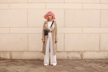 Portrait shooting of a stylish girl with pink hair. Beige shades. Trends of spring and summer 2023.Beige Trench cloak with white top and pants. Black small belt bag