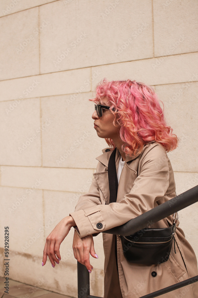 Wall mural Portrait shooting of a stylish girl with pink hair. Beige shades. Trends of spring and summer 2023.Beige Trench cloak with white top and pants. Black small belt bag