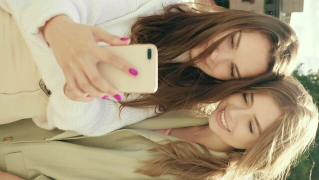 Two young beautiful smiling hipster girls. Sexy carefree women posing in the street . Positive models having fun.They looking at cellphone screen laughing enjoying using mobile apps. Vertical video