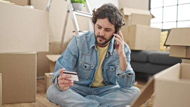 Young hispanic man talking on smartphone holding credit card sitting on floor at new home