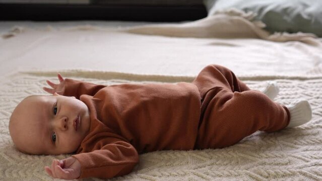 A little boy lies on the bed and looks away. Side view, slow motion. A newborn baby moves his arms. Reflex of early development. The concept of motherhood, family, birth, freedom from swaddling