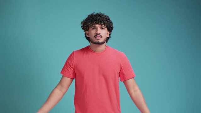 Shocked young bearded Indian man 20s years old says what. wears coral t-shirt on blue studio background. spreading hands shrugging shoulder standing questioned and unaware nothing to say