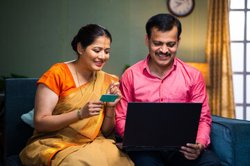 Happy smiling indian couple making online payment for shopping using credit card on laptop on sofa...