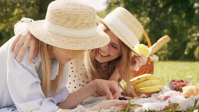 Two young beautiful hipster woman in trendy summer sundress and hats. Carefree women making picnic outside. Positive models sitting on plaid on grass. Reeding book