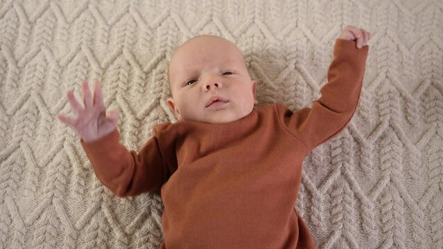 A little boy in a brown sweater lies on a knitted blanket, moves his arms and yawns. Top view, close-up portrait. The concept of motherhood, family, birth, freedom from swaddling. High quality 4k