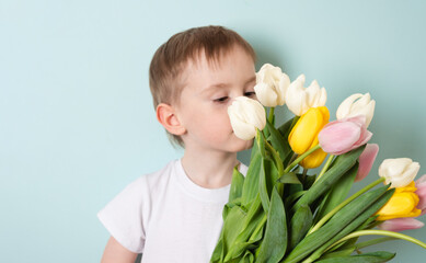 Child boy with freckles in a white T-shirt sniffs fresh bouquet of spring flowers tulips on a blue background.