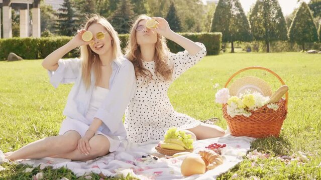 Two young beautiful hipster woman in trendy summer sundress and hats. Carefree women making picnic outside. Positive models sitting on plaid on grass, eating fruits and cheese
