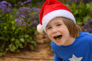 A little boy in a blooming garden in a Santa Claus hat. Concept: Christmas in summer, tropical holidays, traveling with children, snowless winter.