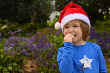 A little boy in a blooming garden in a Santa Claus hat. Concept: Christmas in summer, tropical holidays, traveling with children, snowless winter.