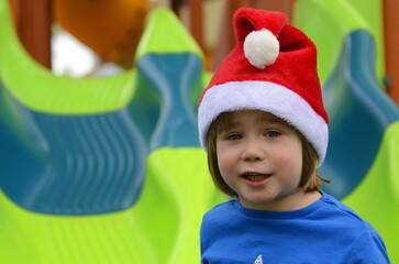 Fototapeta na wymiar Portrait of a little boy in a Santa hat. The child on the playground rides from the children's slide. Concept: Christmas in kindergarten, winter holidays in the tropics. Warm winter