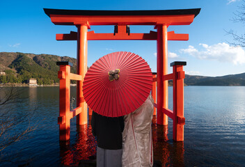 Asian couple in kimono wedding dress standing togather with red umbrella and red torii gate...
