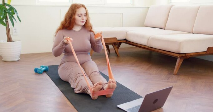 red head obese woman training with resistance band at home. girl with long red hair using flexible belt to do arm stretch and train muscles on yoga mat. lifestyle wellness health and body care
