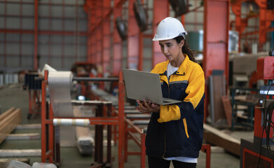 Female industrial engineer in white helmet and safety jacket work in heavy metal engineering factory. Latin technician woman worker using laptop computer in metalwork manufacturing facility, portrait.