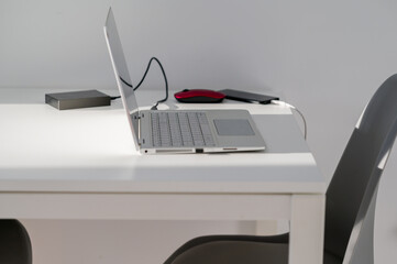 White table with laptop, red mouse, hard drive and phone with gray chairs and white wall. 