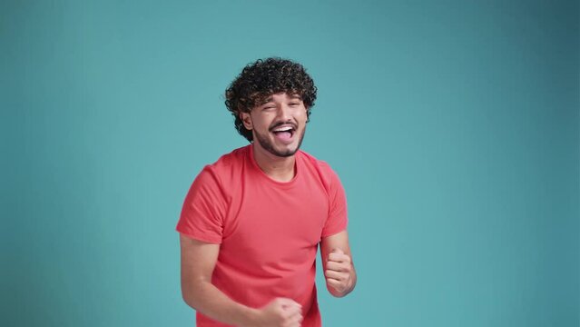 Extremely excited overjoyed man with beard shouting making yes gesture, amazed with his victory, triumph. in coral t-shirt on blue studio background