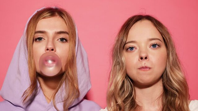 Two young funny smiling playful blond girls dressed in blue hoodie standing and isolated over pink background blowing bubblegum candy bubbles and chewing gum. Women looking at camera