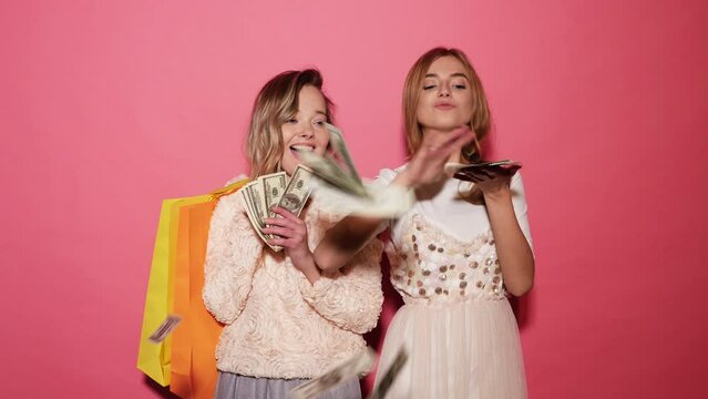 Two young stylish smiling blond women holding shopping bags and making rain money dollars. Hot girls dressed in summer clothes and throwing bills out of a bundle money. Posing on pink background