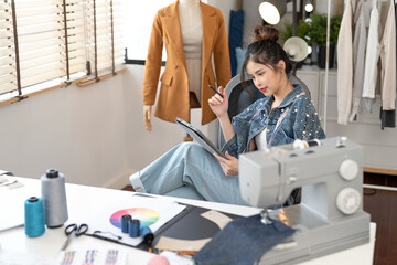 Attractive Asian female fashion designer with measuring tape around her neck siting design dressmaking new clothes collection on laptop in tailoring atelier.