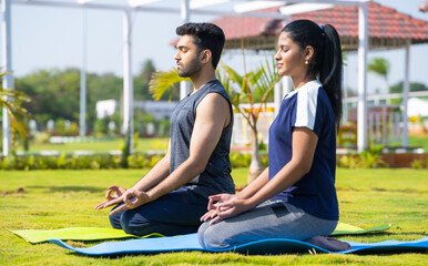 Side view shot of Indian young couple doing yoga or meditation at park during morning - concept of mindfulness, training and tranquility - Powered by Adobe