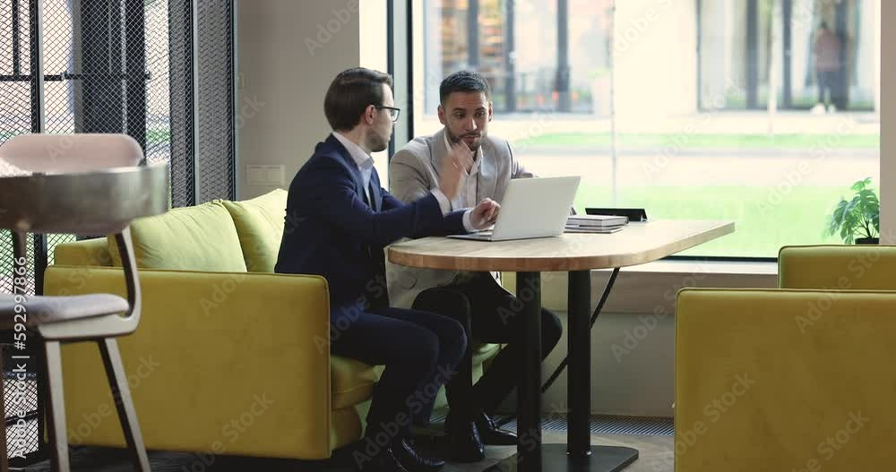 Poster Serious busy business leaders men discussing online project at laptop in office cafe, sitting at table, talking, speaking, using computer, watching content, negotiating on work issues