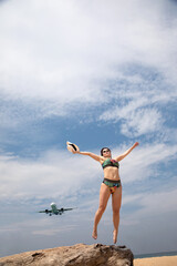 A beautiful girl poses on the beach against the backdrop of a landing plane. Sandy beach. Sunny day. Big plane.