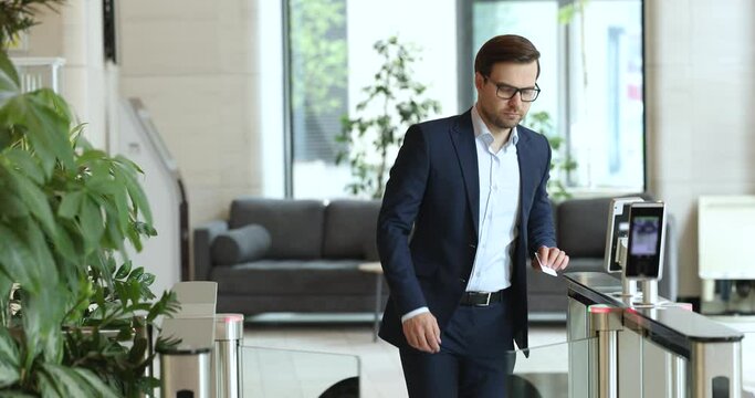 Handsome businessman in formal suit and glasses going in business center building, walking across hall, applying keycard to sensor control keypad, walking pass camera with serious face