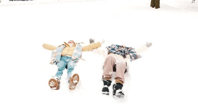Two young beautiful smiling hipster female in trendy warm clothes and scarfs. Carefree women posing in the street in park. Positive pure models hugging each other. They lying in snow. Going crazy
