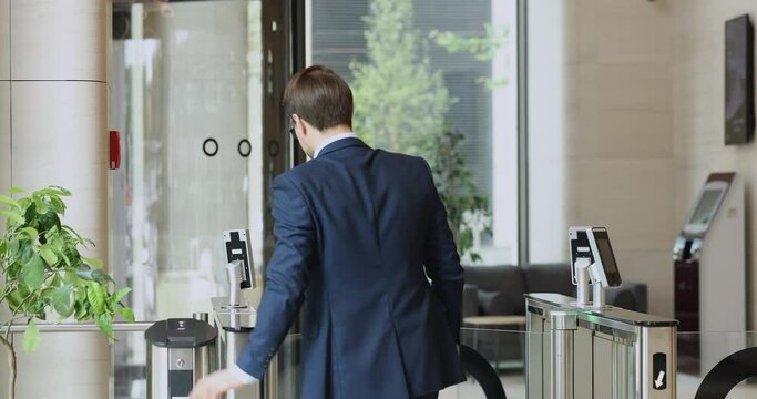 Young businessman in formal suit using electronic keycard, leaving office building, applying identity badge to sensor for gate opening at entrance, walking through business center security equipment
