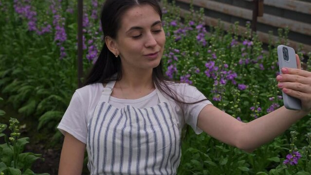 a girl in a striped apron, smiling and posing, takes a selfie on her smartphone against the background of flower rows with matiola. Homemade homemade greenhouse. Family business and business