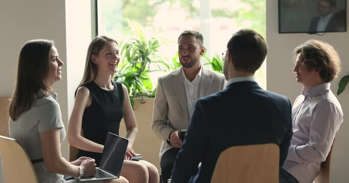 Happy business colleagues sitting in circle in office, brainstorming on project ideas, talking, listening to speaker woman, laughing, enjoying professional networking on group teambuilding meeting