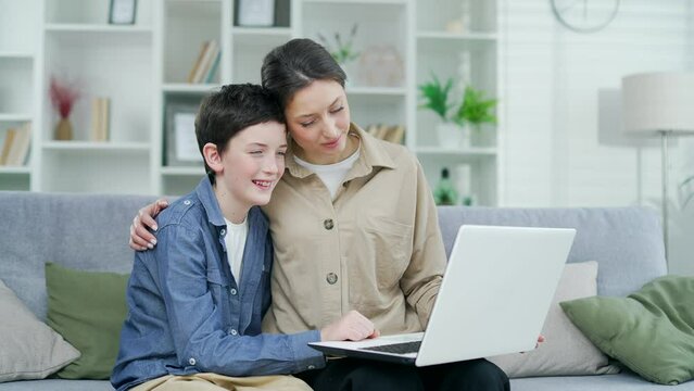Mother and son hugging together sitting on sofa at home in living room watching video online using laptop, family spending time together watching online educational course