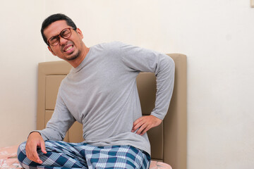 Adult Asian man sitting on his bed suffering lower back pain - obrazy, fototapety, plakaty