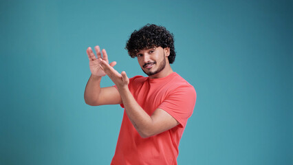 Young handsome Indian man with beard in coral t-shirt on blue studio background doing money gesture...
