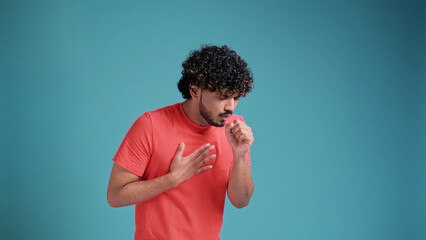 Young indian man in coral t-shirt on blue studio background, scarf sneezing or coughing covering...