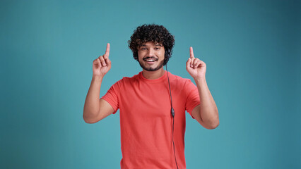 Young indian call center agent man using headset in coral t-shirt on blue studio backgroun pointing...
