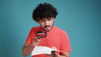 Funny Indian man with magnifying glass is reading book in coral t-shirt on blue studio background