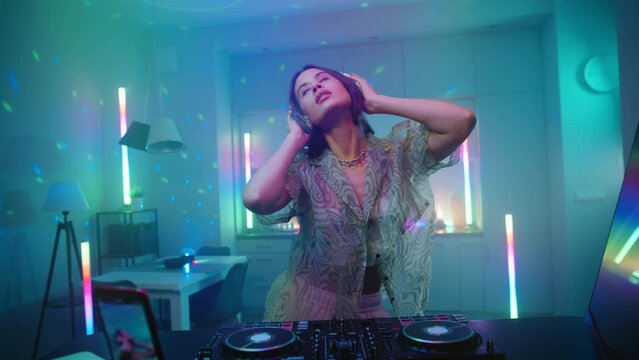 Cheerful young woman listening music dancing at home create modern music at turntables, cute female dj streaming podcast enjoying music set in apartment lit with colorful glowing neon light