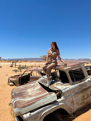 Young woman in safari overalls sits on the rooftop of abandoned old rusty car.