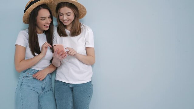 Two young beautiful smiling hipster female in trendy summer  t-shirt and jeans clothes. Sexy carefree women posing near wall in studio.Positive models looking at smartphone screen. Using apps 