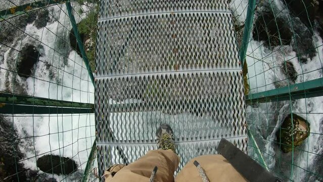 First person view looking down at is feet while crossing a metal bridge over a powerfull river.
