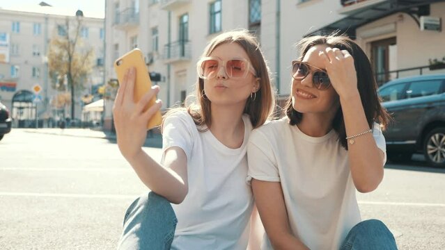 Two young beautiful smiling hipster female in trendy summer white t-shirt clothes and jeans.Sexy carefree women posing on the street background. Positive models having fun, hugging and take selfie 
